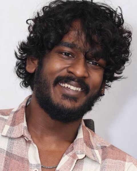 Aegan smiling with curly hair and a beard in a pink checked shirt, head-and-shoulders portrait