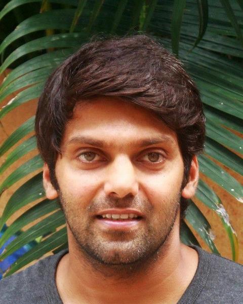 Arya smiling in a grey T-shirt in front of green palm leaves