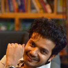 Sreenath Bhasi smiling with his hand raised, seated portrait