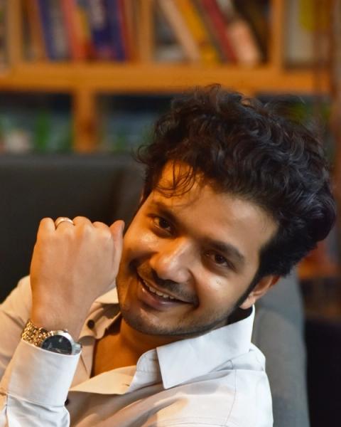 Sreenath Bhasi smiling with his hand raised, seated portrait