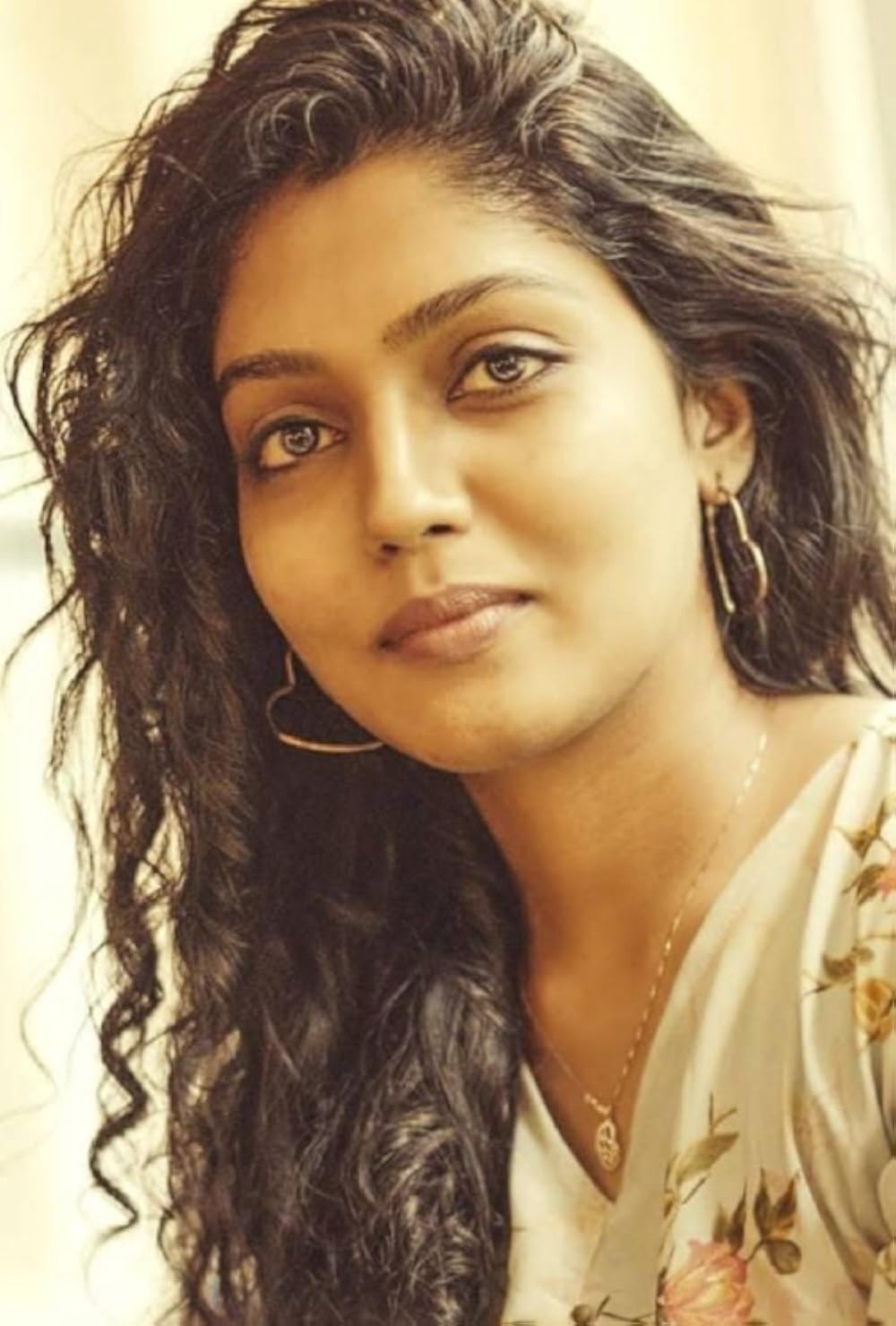 Chaithra Praveen with long curly hair, hoop earrings and a floral top, looking at the camera