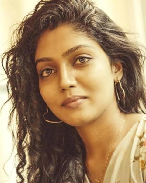 Chaithra Praveen with long curly hair, hoop earrings and a floral top, looking at the camera
