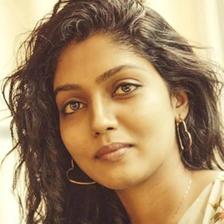 Chaithra Praveen with long curly hair, hoop earrings and a floral top, looking at the camera