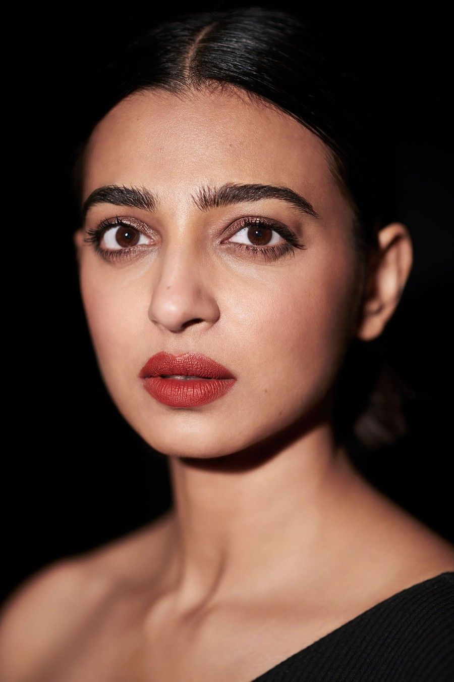 Radhika Apte with her hair pulled back and red lipstick against a black background, portrait