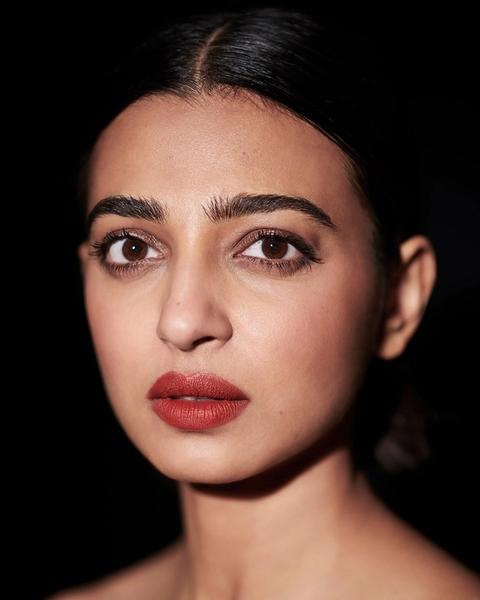 Radhika Apte with her hair pulled back and red lipstick against a black background, portrait