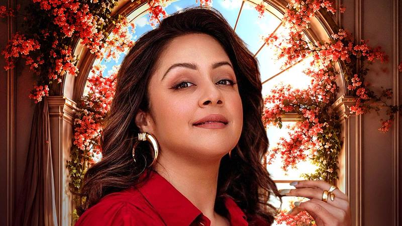 Jyothika smiling in a red outfit under a floral arch