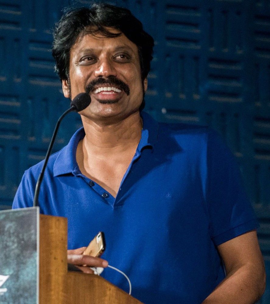 S. J. Suryah speaking at a podium in a blue polo shirt
