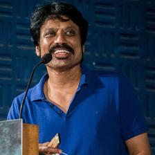 S. J. Suryah speaking at a podium in a blue polo shirt