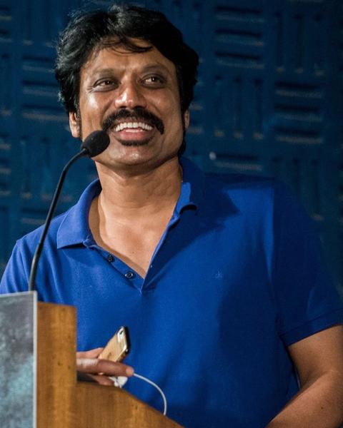 S. J. Suryah speaking at a podium in a blue polo shirt
