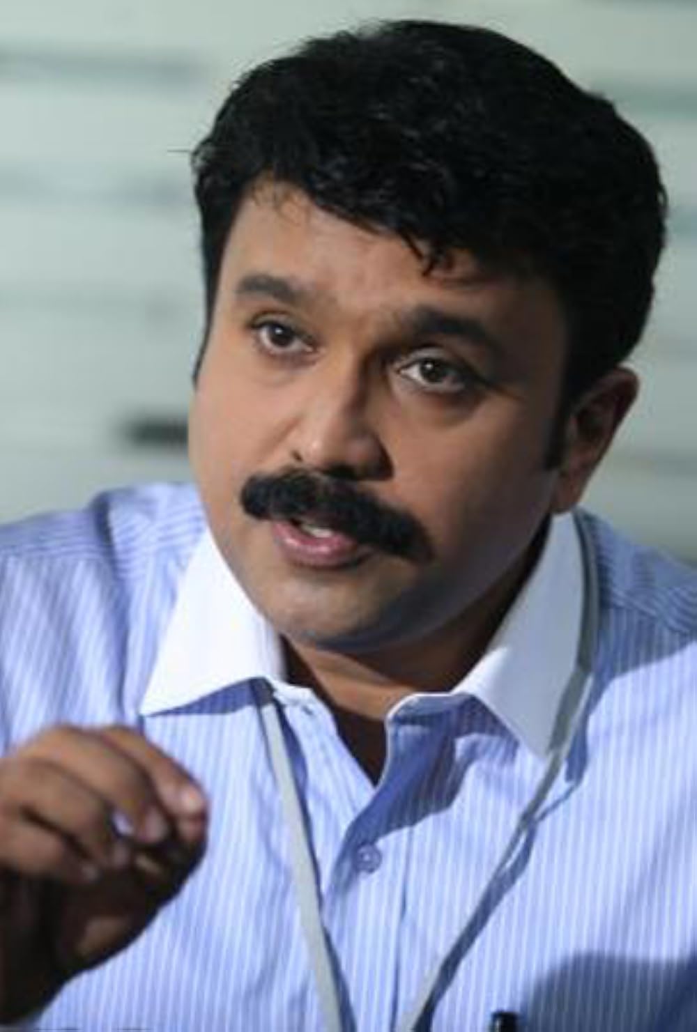 Sudheesh with a moustache, wearing a blue striped shirt, speaking and gesturing with one hand