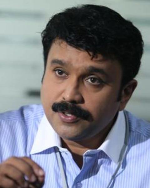 Sudheesh with a moustache, wearing a blue striped shirt, speaking and gesturing with one hand