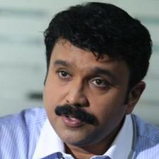 Sudheesh with a moustache, wearing a blue striped shirt, speaking and gesturing with one hand