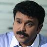 Sudheesh with a moustache, wearing a blue striped shirt, speaking and gesturing with one hand