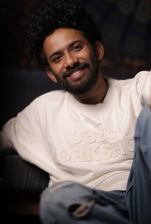 Christo Xavier smiling with curly hair and a beard, in a white sweatshirt and blue jeans, seated portrait