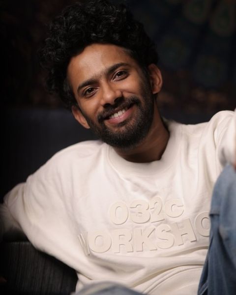 Christo Xavier smiling with curly hair and a beard, in a white sweatshirt and blue jeans, seated portrait