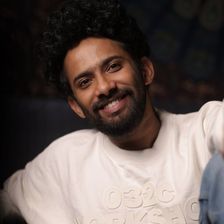 Christo Xavier smiling with curly hair and a beard, in a white sweatshirt and blue jeans, seated portrait