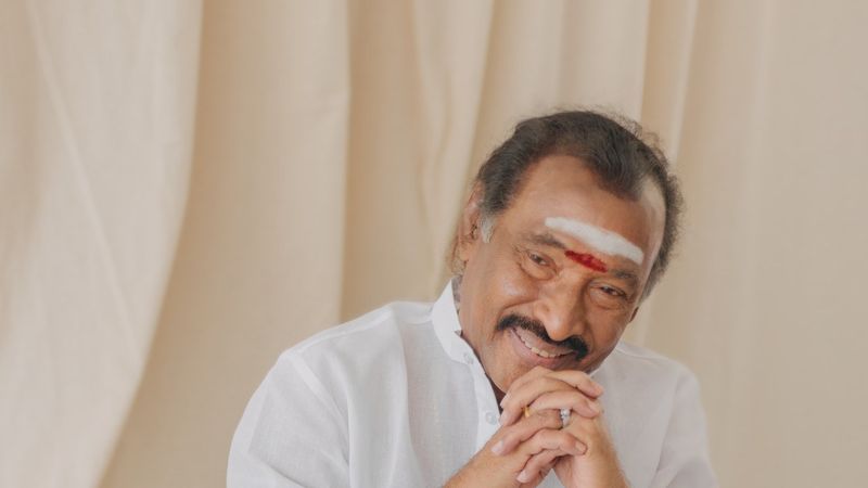 Composer Deva smiling with his hands clasped, in a white shirt against a cream curtain