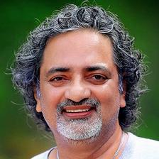 Joy Mathew with grey curly hair and a white beard in a white T-shirt, smiling