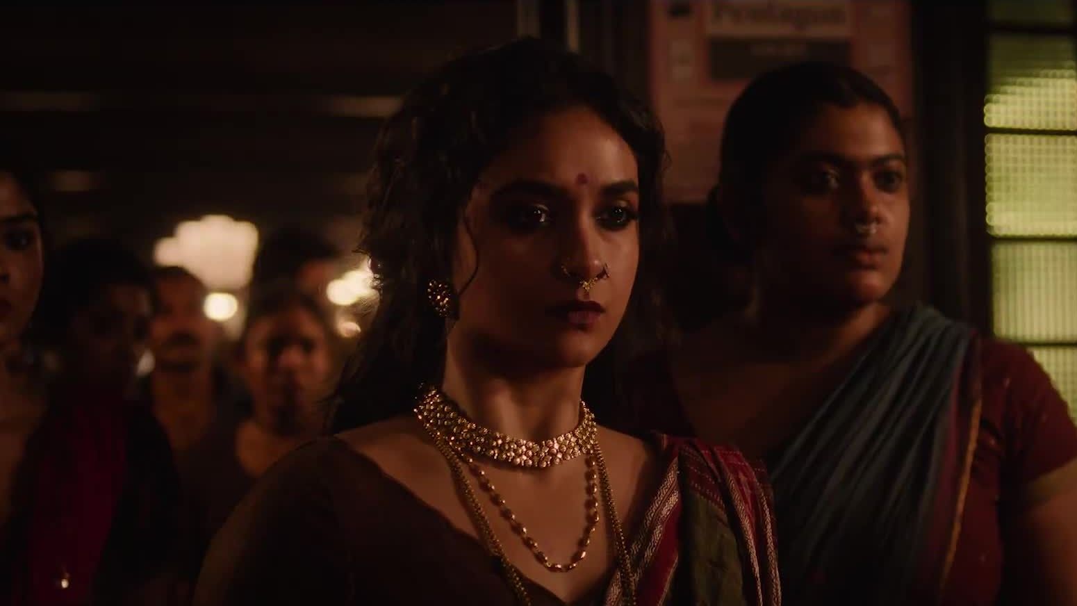 Keerthy Suresh in a gold choker and sari stands among women in a dim hall in a still from the Akka teaser