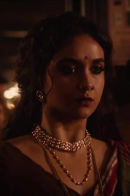 Keerthy Suresh in a gold choker and sari stands among women in a dim hall in a still from the Akka teaser