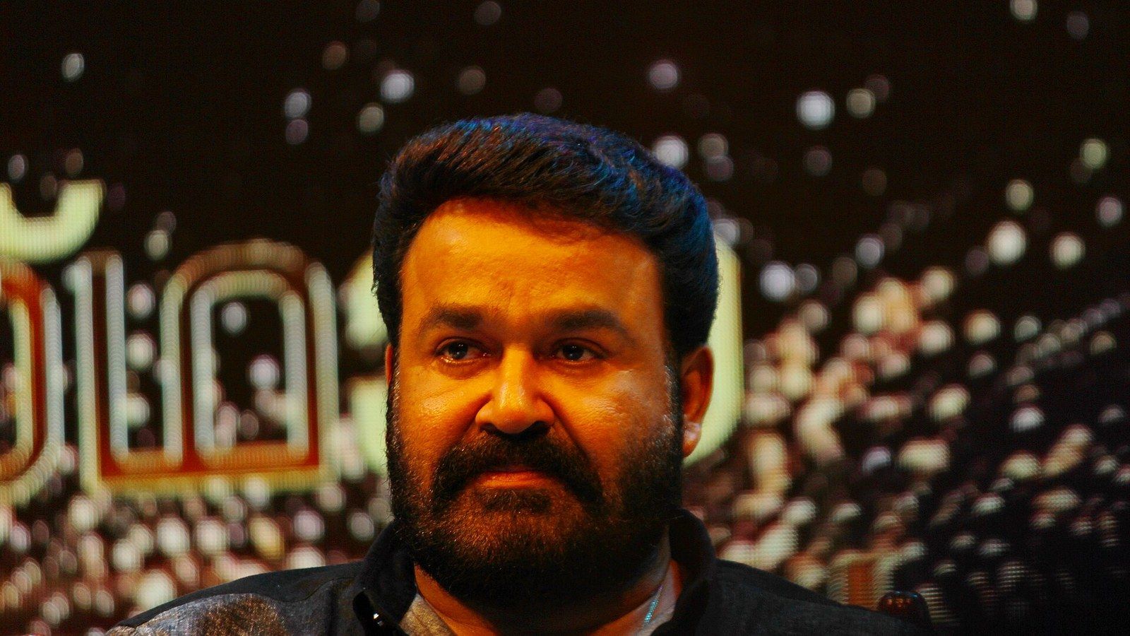 Mohanlal, in a dark shirt with a full beard, looks on intently at an event.