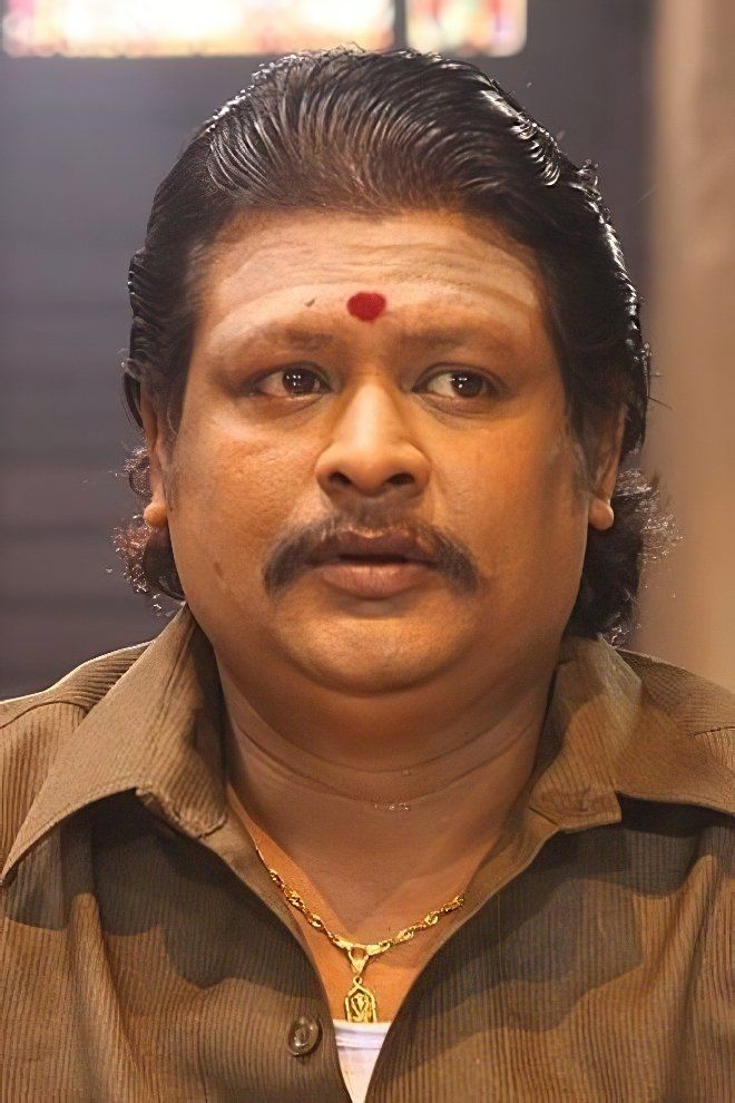 Namo Narayana with slicked-back hair, a red bindi and a brown shirt, looking ahead