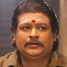 Namo Narayana with slicked-back hair, a red bindi and a brown shirt, looking ahead