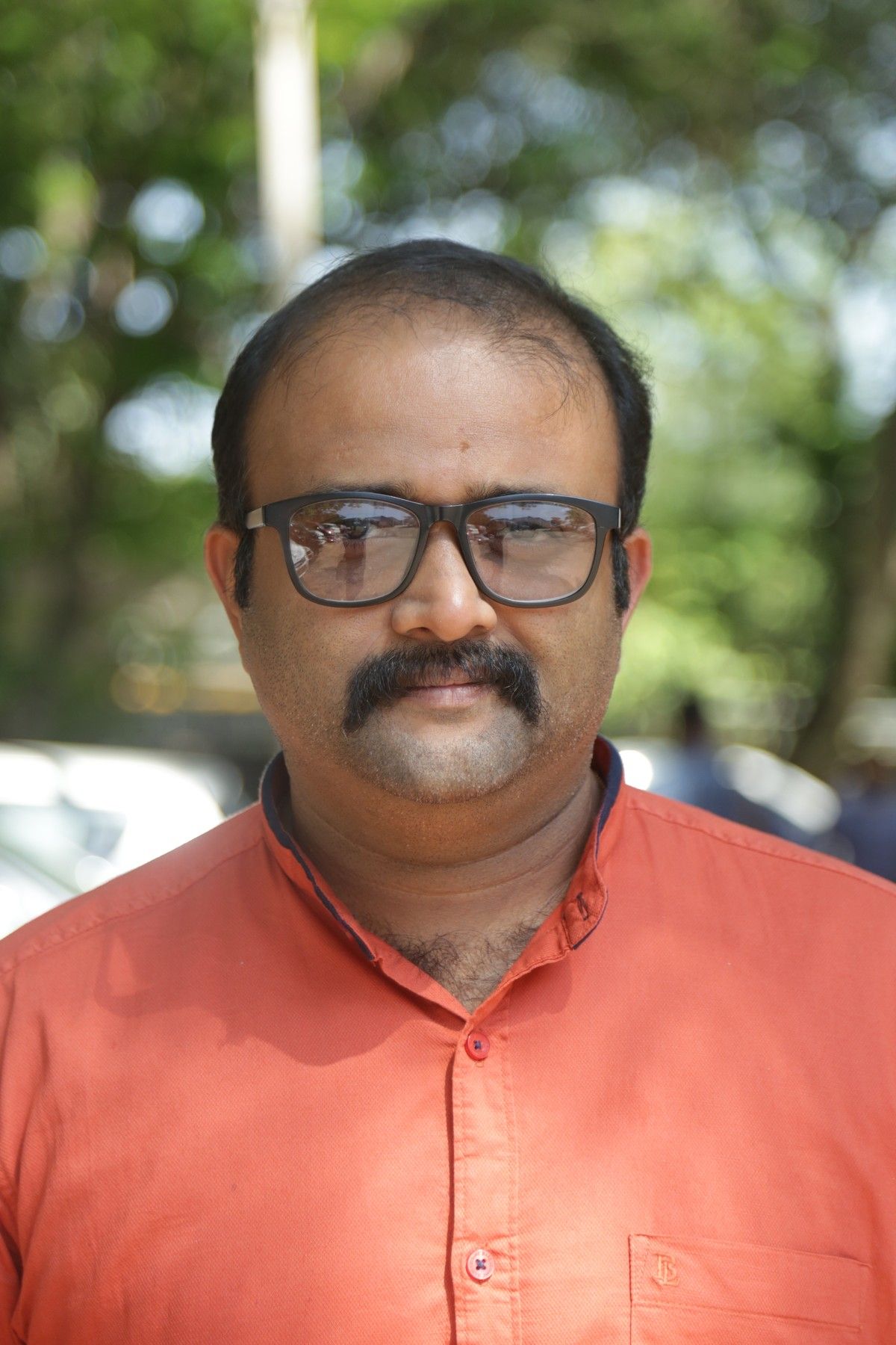 Alexander Prasanth in black-framed glasses and an orange shirt, standing outdoors under trees