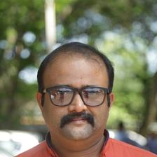 Alexander Prasanth in black-framed glasses and an orange shirt, standing outdoors under trees