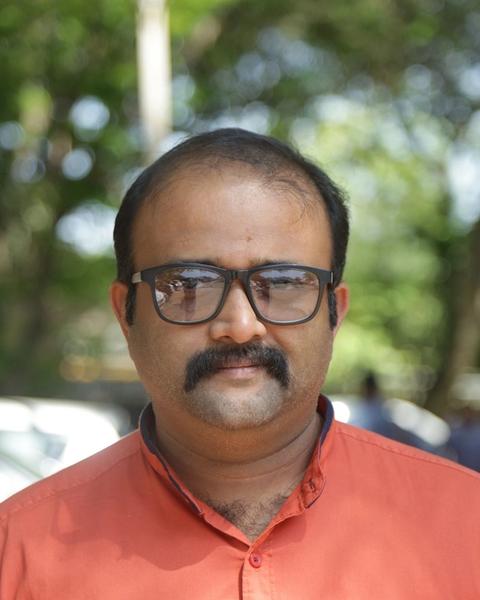 Alexander Prasanth in black-framed glasses and an orange shirt, standing outdoors under trees