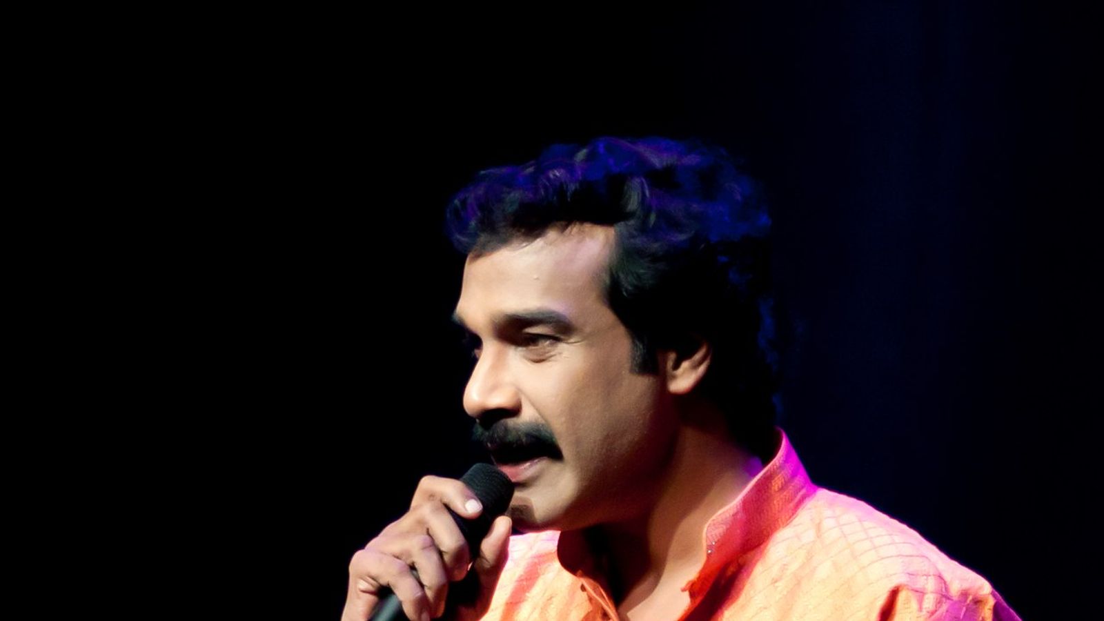 Jayaraj Warrier in an orange kurta speaking into a microphone on a dark stage.