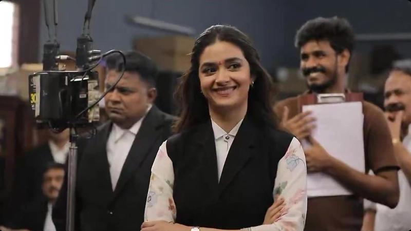 Keerthy Suresh smiling in a lawyer's black vest on the Sathyavan Savithiri set, with crew members holding a clapboard and a camera rig beside her