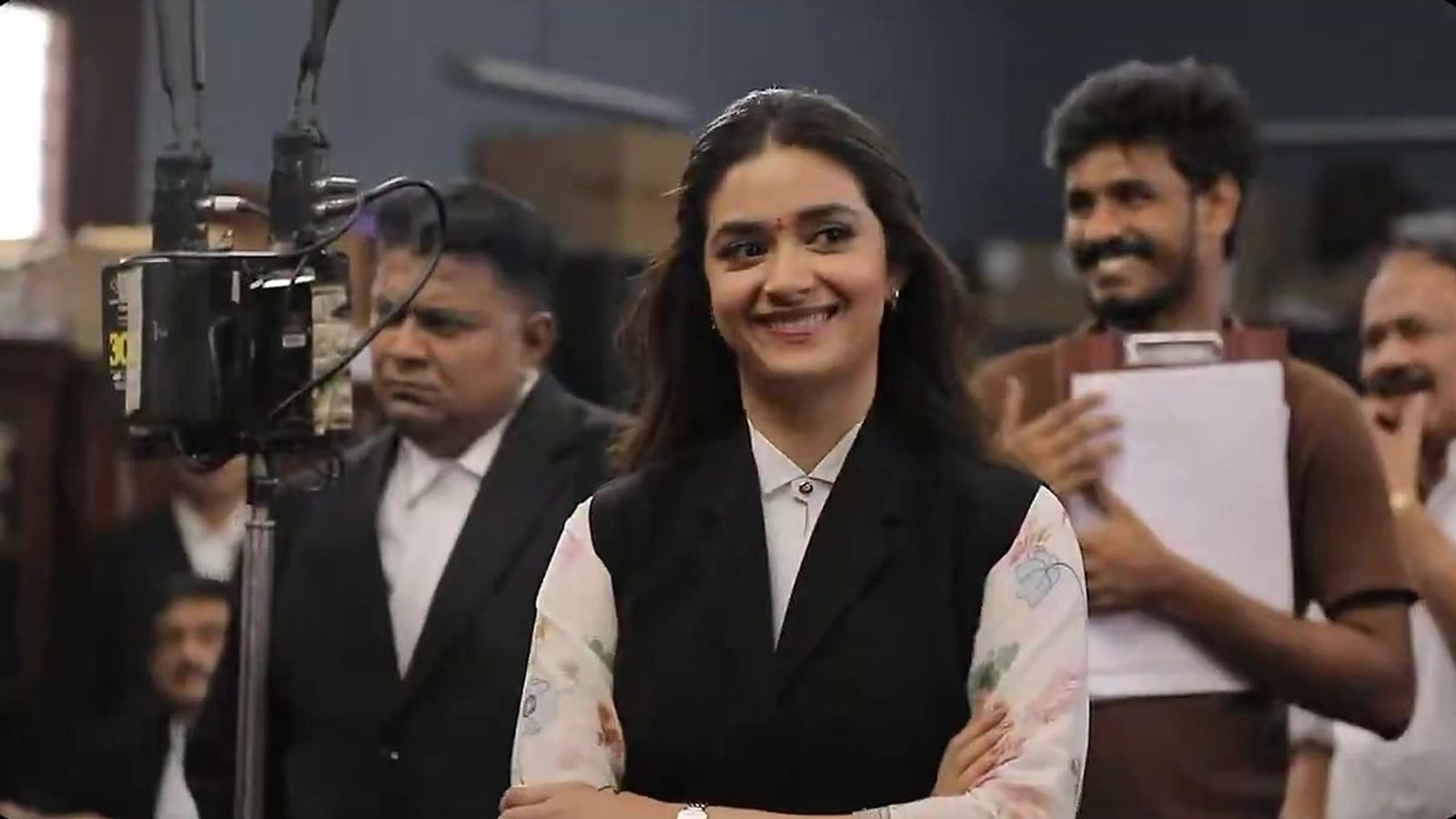 Keerthy Suresh smiling in a lawyer's black vest on the Sathyavan Savithiri set, with crew members holding a clapboard and a camera rig beside her