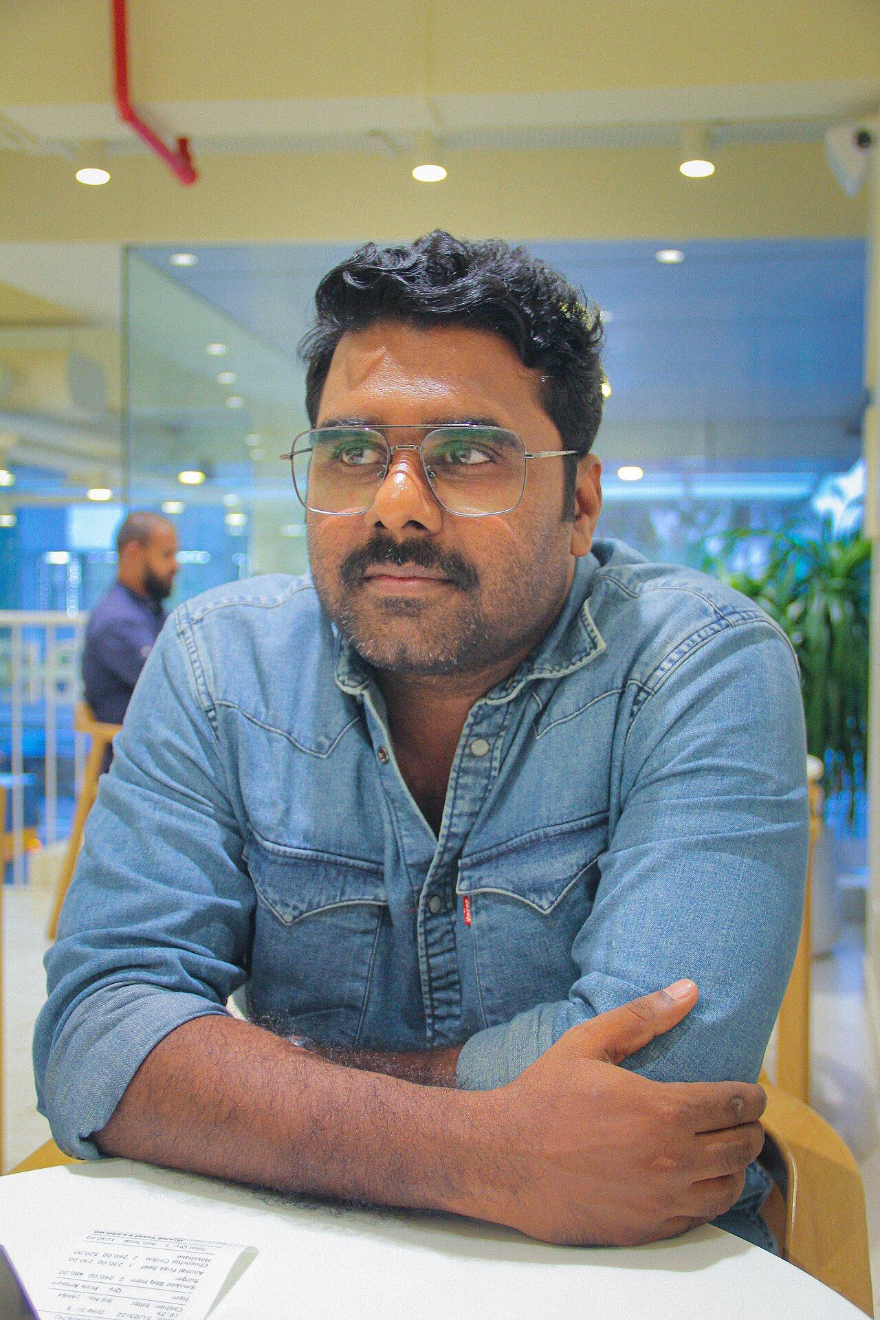 Sajin Cherukayil in glasses and a denim shirt, seated at a table