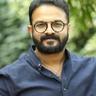 Jayasurya in round glasses and a blue shirt outdoors, portrait