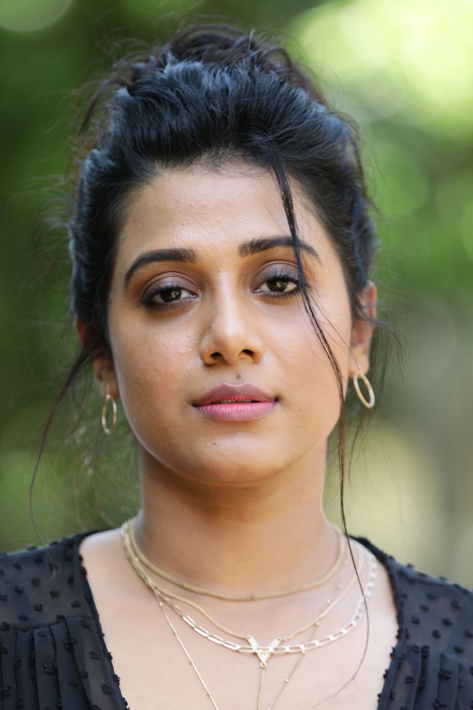 Shilpa Manjunath with her hair tied up, wearing a black top and layered necklaces against green foliage, portrait