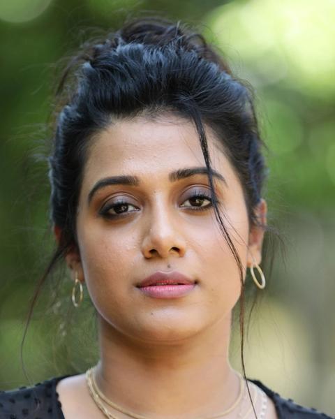 Shilpa Manjunath with her hair tied up, wearing a black top and layered necklaces against green foliage, portrait