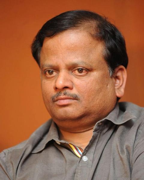 K. V. Anand in a grey shirt against an orange background