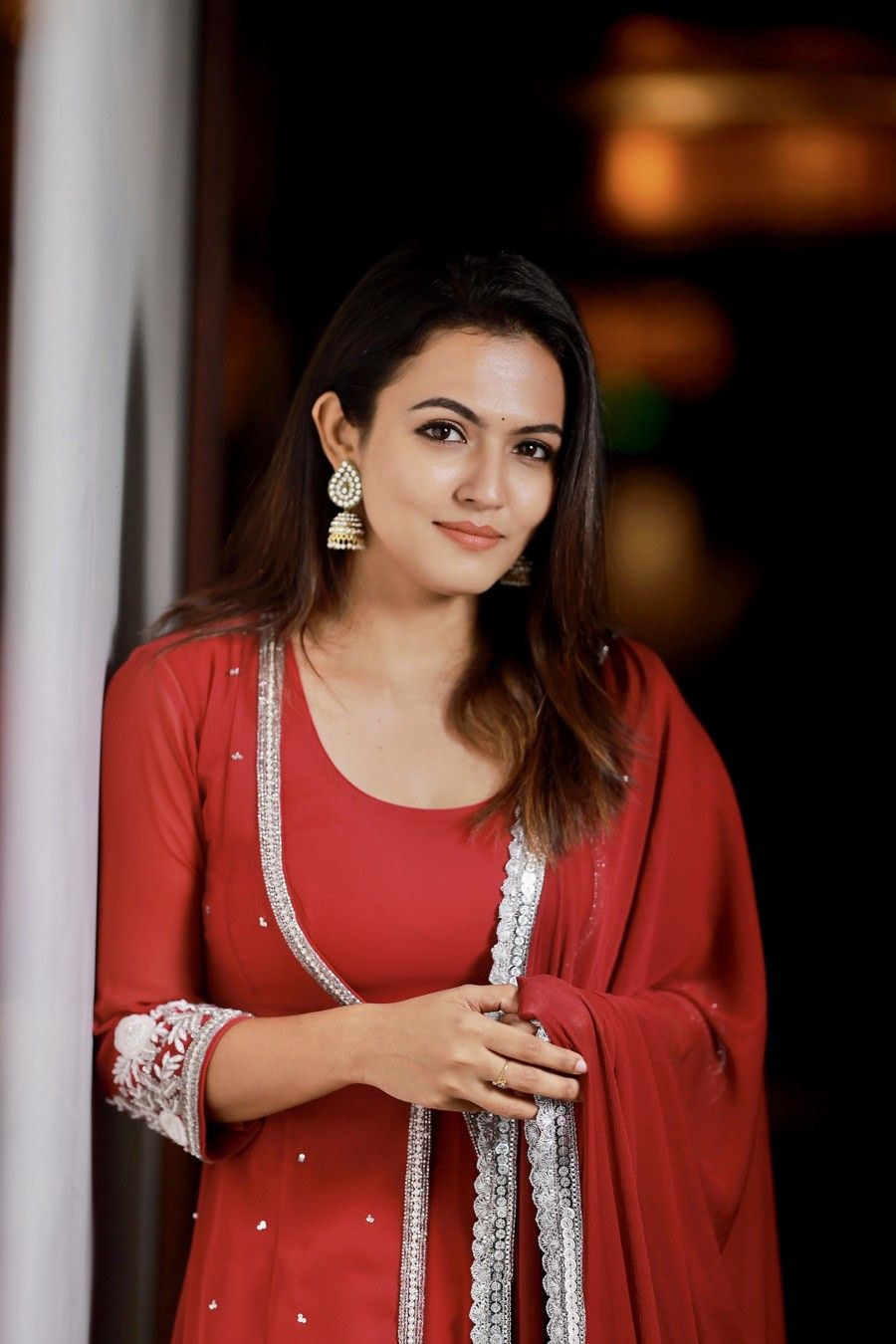 Aparna Das in a red outfit with a white dupatta, leaning against a pillar