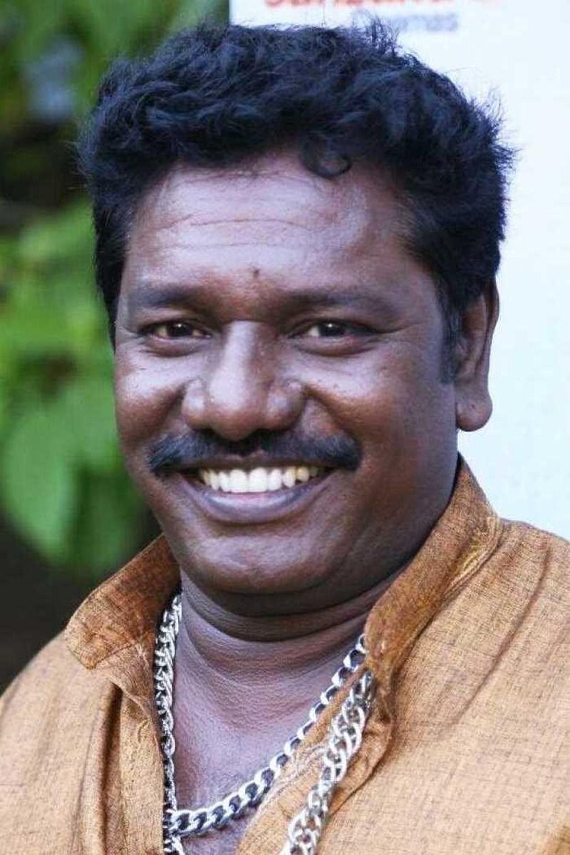 Karunas smiling widely with a moustache and gold chain, close-up