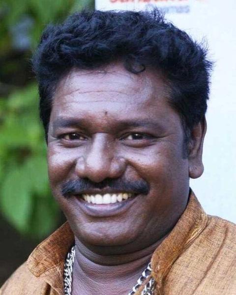 Karunas smiling widely with a moustache and gold chain, close-up