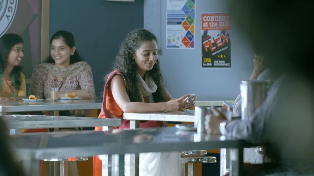 Darshana Rajendran smiling at her phone in a canteen scene from the Hridayam trailer