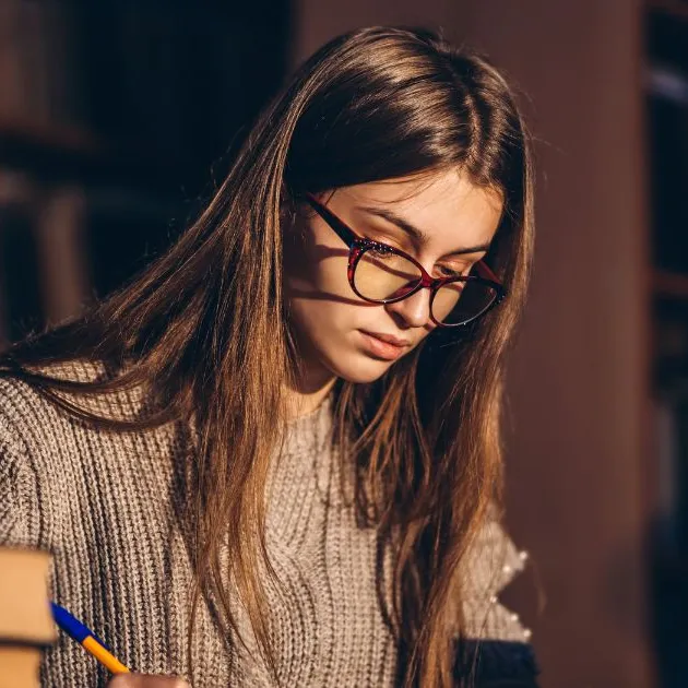 A focused student wearing glasses and studying in a library, surrounded by books, symbolizing dedication to learning and research.