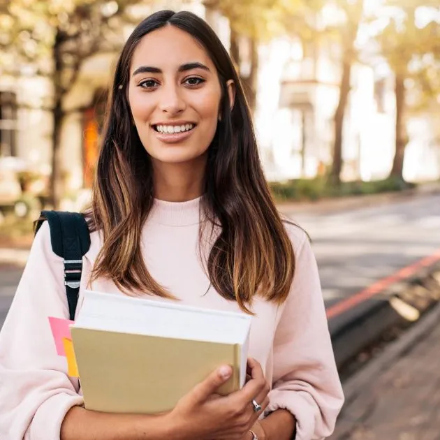 A smiling student holding books, standing on a sunny tree-lined street, symbolizing academic guidance and preparation.