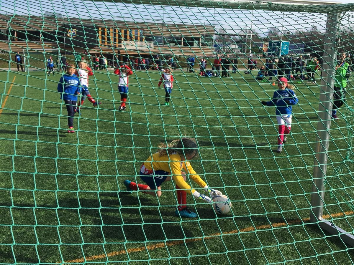 Ung keeper i gul drakt plukker opp ballen foran målet, sett gjennom nettmaskene bak mål