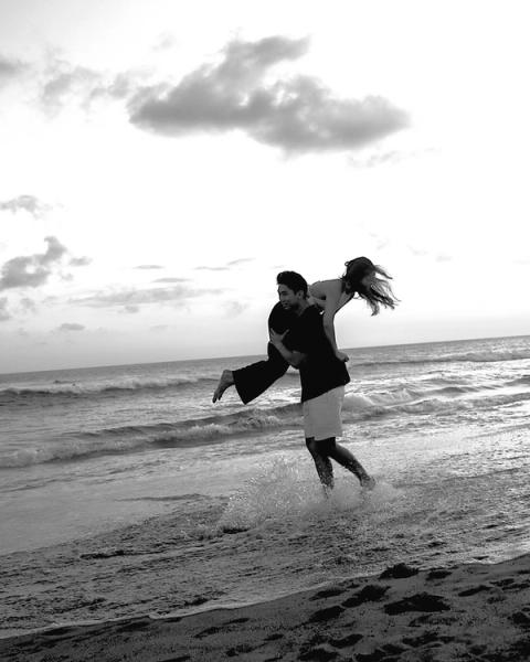 Black and white photo of a man carrying his partner piggyback through the surf