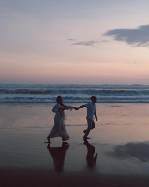 Woman in a flowing white dress holding a bouquet, running hand in hand with her partner along the shore at dusk