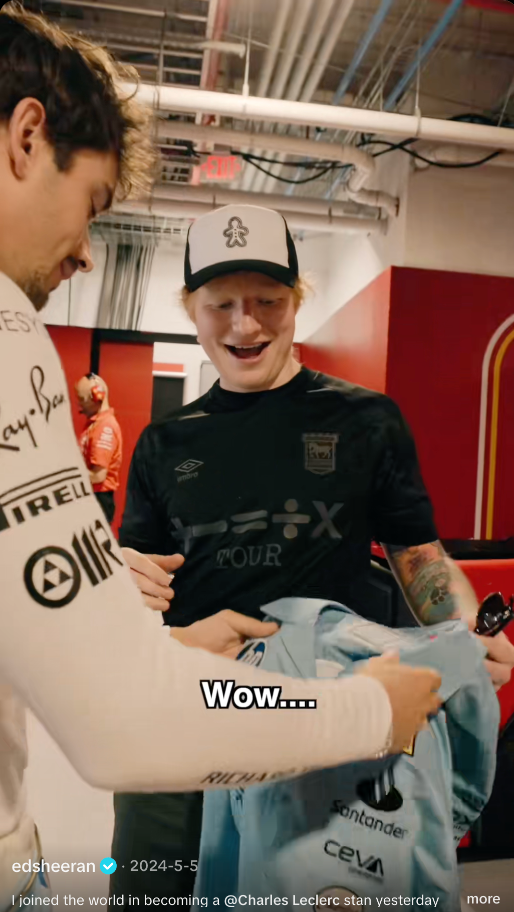 Ed Sheeran, wearing a black shirt and a cap with a gingerbread man, smiles as he is handed a light blue jersey.