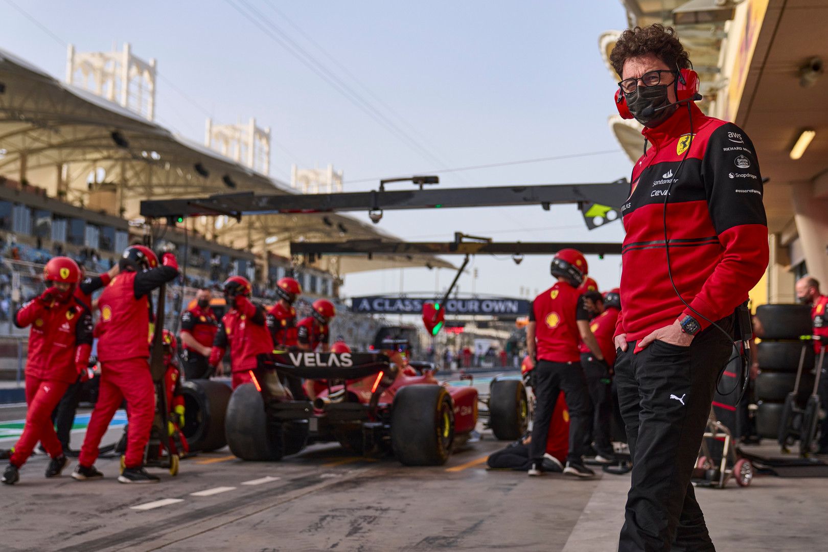 A man in a Ferrari team uniform watches a pit crew perform a pit stop on a red F1 car.