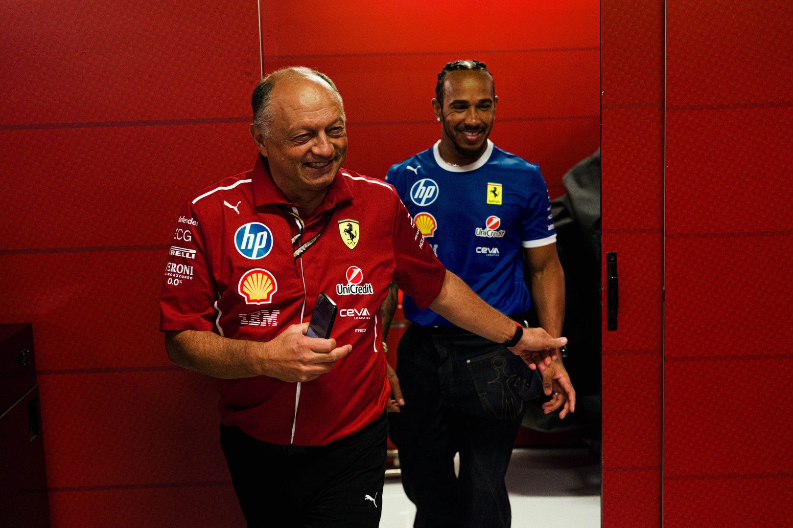 Lewis Hamilton in a blue shirt smiles behind an older man in a red Ferrari team shirt, both in a red hallway.